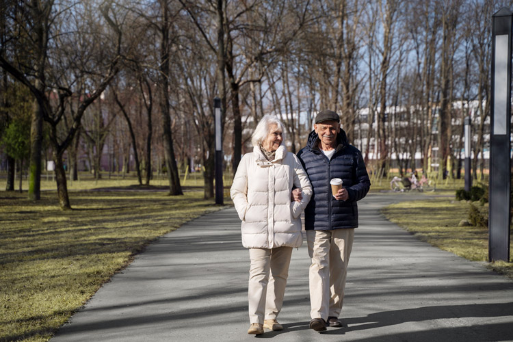 An older couple taking a walk in the park. The man is holding a cup of coffee.